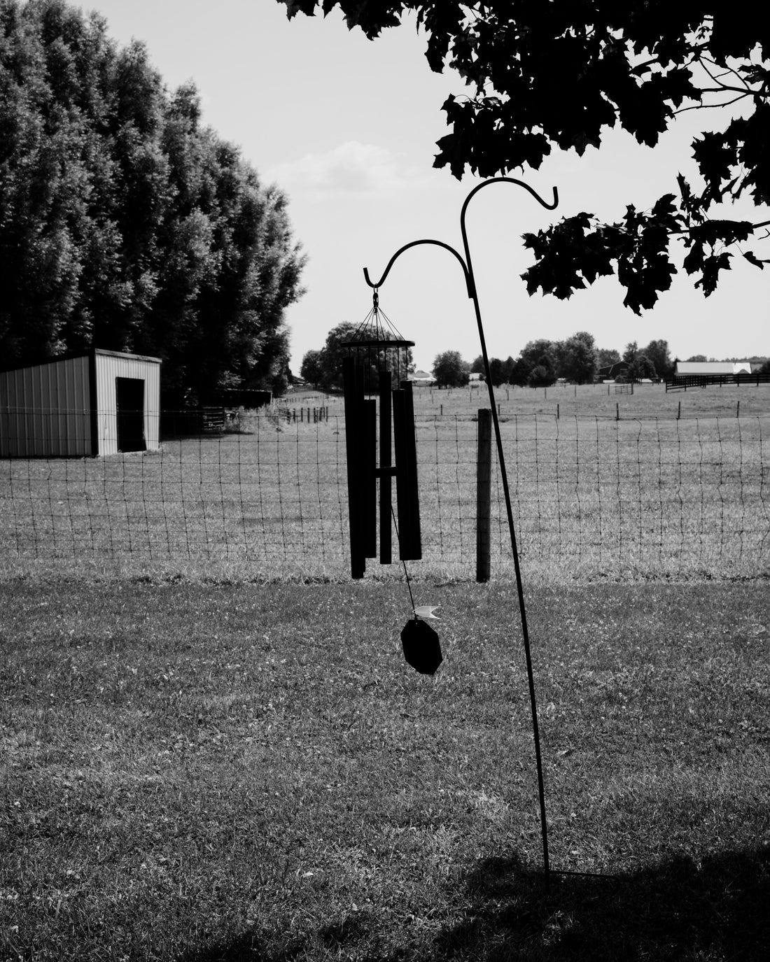 Black and white photo of an Amish handcrafted wind chime hanging from a shepherd's hook garden stake in a rural Indiana field with a barn in the background