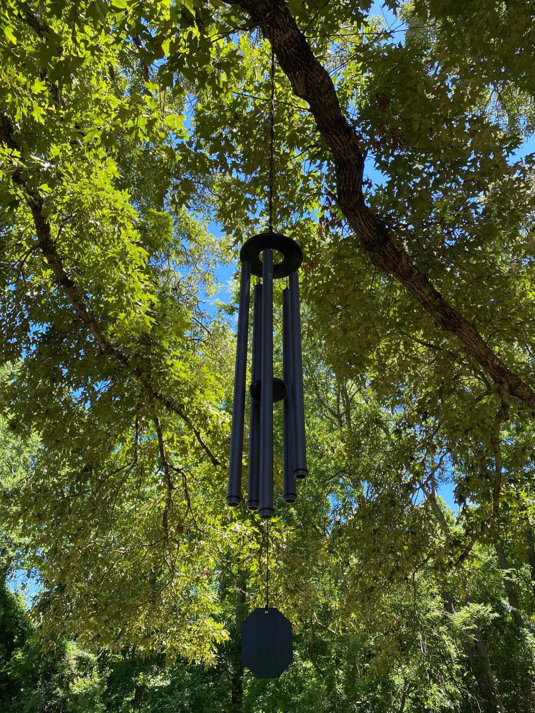 Bronze Amish handcrafted wind chime hanging from a tree branch surrounded by lush green summer leaves with blue sky peeking through