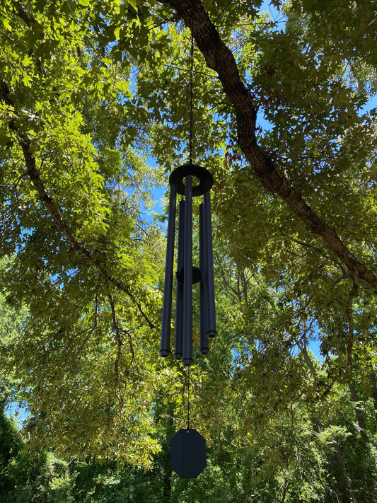 Bronze Amish handcrafted wind chime hanging from a tree branch surrounded by lush green summer leaves with blue sky peeking through