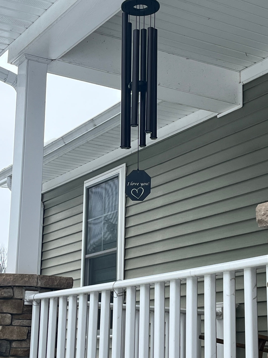 Black wind chime with custom engraved sail reading I love you with a heart, hanging from a covered porch ceiling with white railing and stone pillars