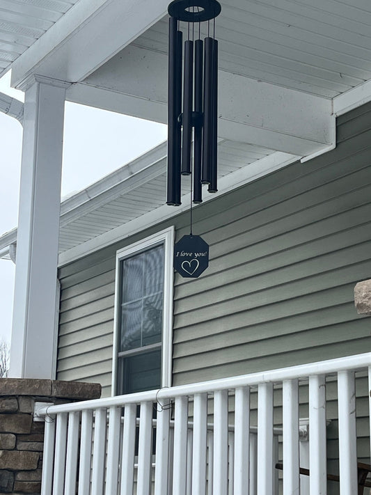 Black wind chime with custom engraved sail reading I love you with a heart, hanging from a covered porch ceiling with white railing and stone pillars