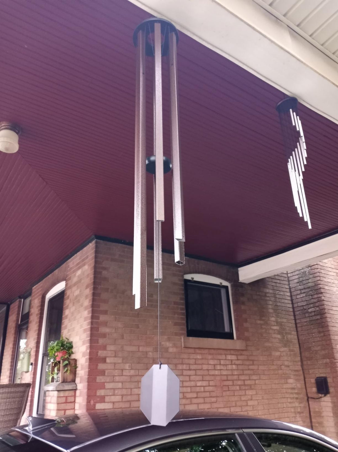 Two wind chimes hanging from a burgundy beadboard porch ceiling, a large silver aluminum chime in the foreground and a smaller chime in the background, brick house