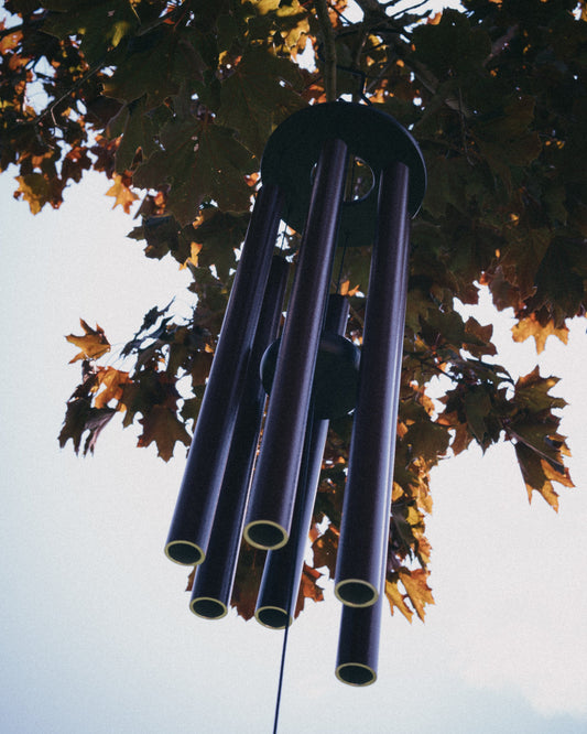 Looking up at six aluminum wind chime tubes of varying lengths hanging from a round top plate, surrounded by autumn maple leaves against the sky