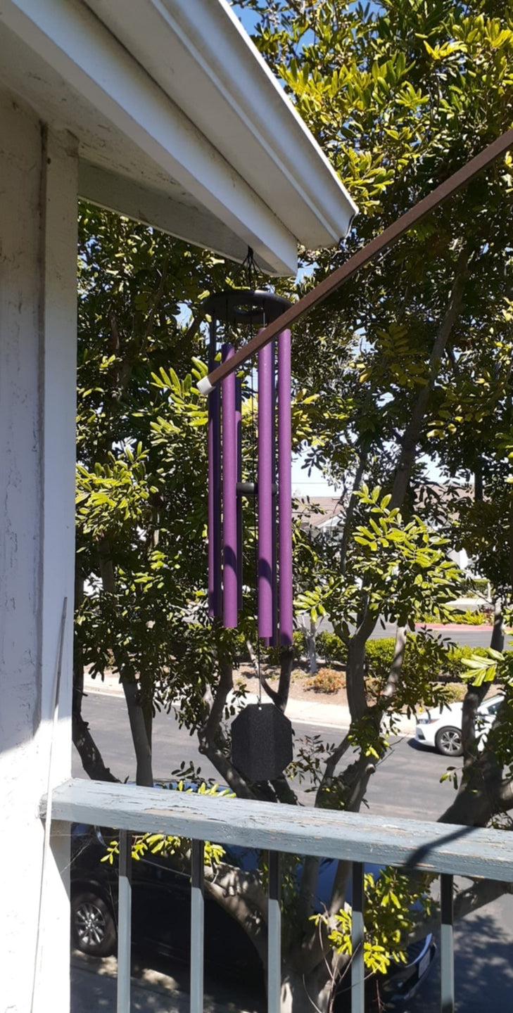 Purple Enchanting Flowers wind chime hanging from a porch eave with green trees and a sunny neighborhood visible in the background