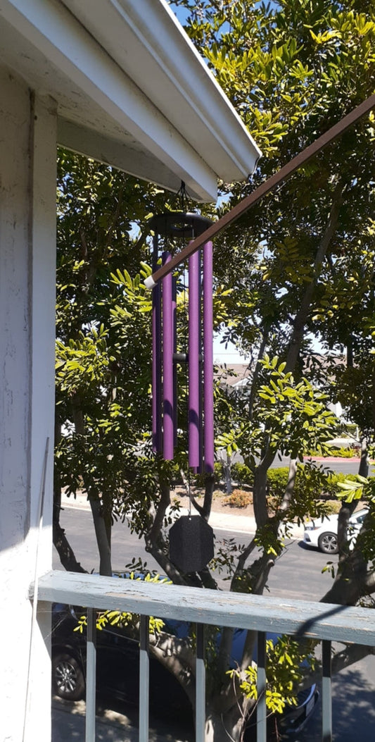 Purple Enchanting Flowers wind chime hanging from a porch eave with green trees and a sunny neighborhood visible in the background