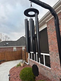 Large black deep tone wind chime hanging from a heavy duty bracket on a brick house with an overcast sky in the background