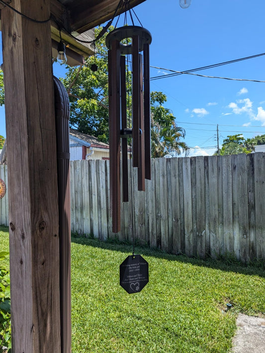 Pet memorial wind chime with custom engraved sail reading In loving memory with paw print graphics, brown finish, hanging from a backyard pergola post