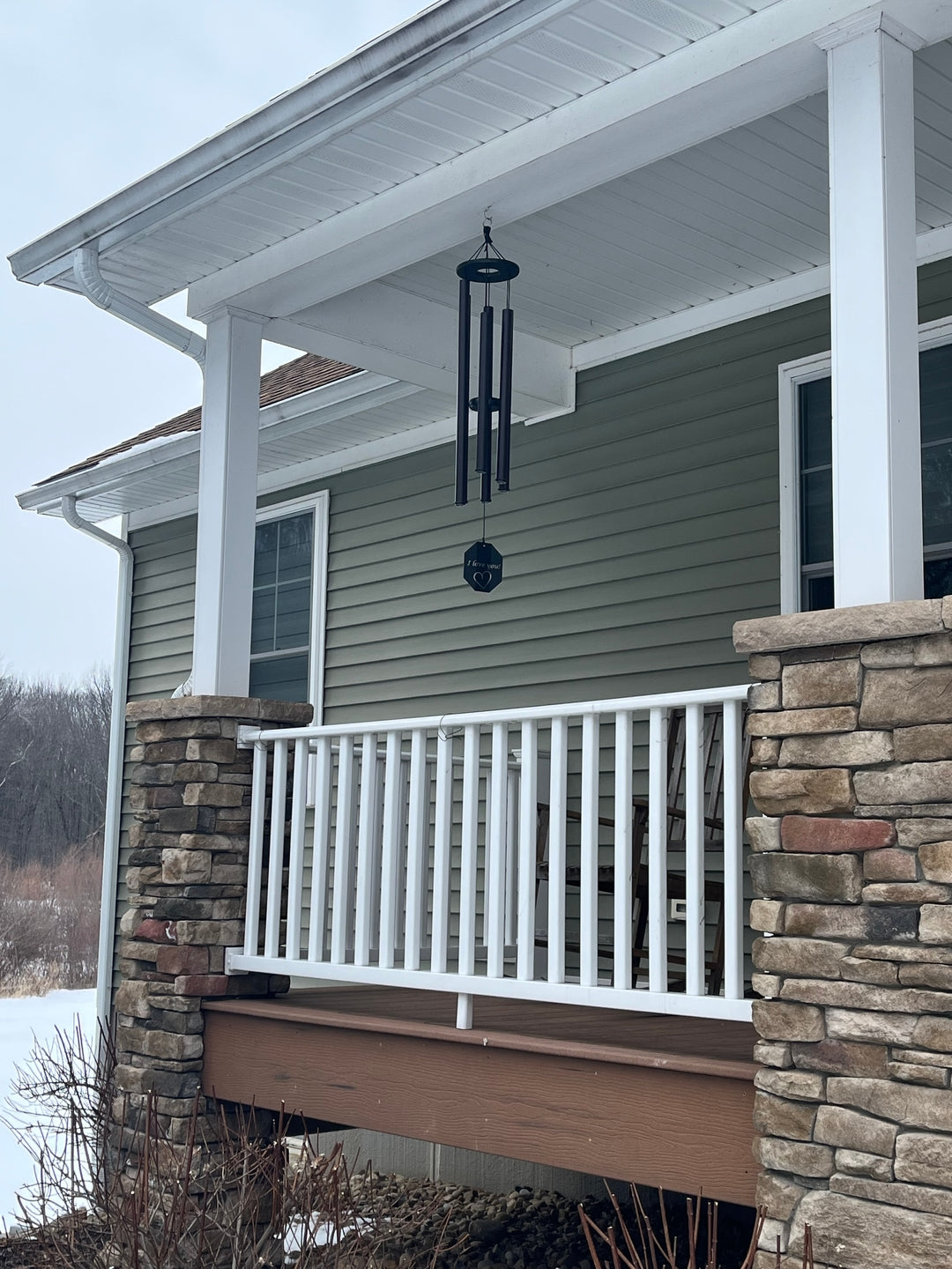 Engraved wind chime hanging from a covered porch in winter with snow on the ground, stone pillars, white railing, and sage green siding