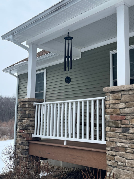 Engraved wind chime hanging from a covered porch in winter with snow on the ground, stone pillars, white railing, and sage green siding