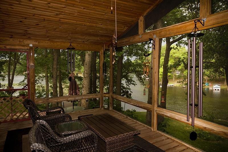 Screened-in lakehouse porch with multiple wind chimes hanging from the ceiling, wicker furniture, and a lake view through the trees